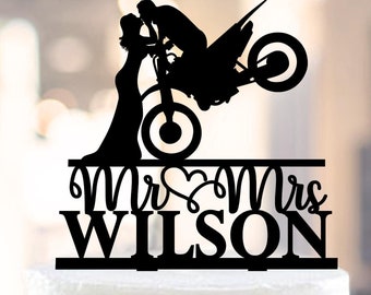 May include: Black silhouette of a couple kissing on a dirt bike, with the words "Mr & Mrs WILSON" below. This is a cake topper.