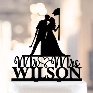Op de afbeelding: Zwarte silhouet van een stel dat elkaar kust, met een golfclub en een vlag, met de tekst "Mr & Mrs WILSON" eronder. Dit is een taarttopper.
