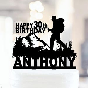 以下が含まれることがあります： 「HAPPY 30th BIRTHDAY ANTHONY」の文字と、バックパック、トレッキングポール、山の風景を持つハイカーのシルエットが描かれた黒いケーキトッパー。白いケーキの上にトッパーが置かれています。
