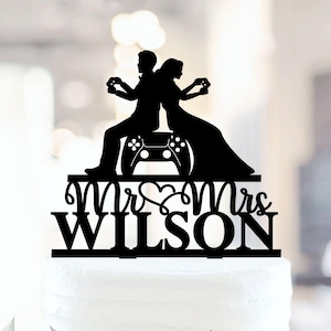 May include: Black cake topper featuring a silhouette of a couple, back-to-back, holding game controllers. A large game controller is between them. The words "Mr & Mrs WILSON" are below the couple. The topper is on a white cake.