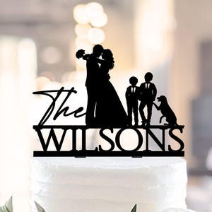 May include: Black silhouette cake topper depicting a bride and groom, two children, and a dog. The text "The Wilsons" is displayed below the figures. The topper is placed on a white cake with greenery.