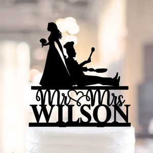 Può includere: Topper per torta in silhouette nera di una sposa e uno sposo. Lo sposo tiene una spatola e una padella. Il testo "Mr & Mrs WILSON" è sotto le figure.