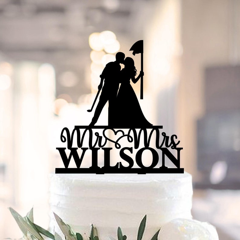 Op de afbeelding: Zwarte silhouet van een stel dat elkaar kust, met een golfclub en een vlag, met de tekst "Mr & Mrs WILSON" eronder. Dit is een taarttopper.
