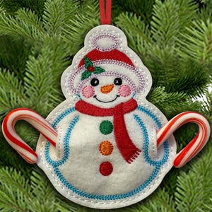 May include: A snowman-shaped Christmas ornament with a red scarf and hat, holding two candy canes. The ornament is white with embroidered details, including a smiling face, and is trimmed with blue stitching. A red ribbon is attached for hanging.