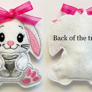May include: A white felt bunny treat bag with pink ears, a pink bow, and a clear plastic window for holding treats. The back of the bag is plain white felt. "Back of the treat bag" is written on the back of the bag.