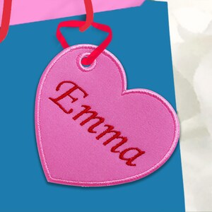 Puede incluir: Una etiqueta rosa en forma de corazón con el nombre "Emma" bordado en escritura roja. La etiqueta está unida a una bolsa de regalo azul con asas de cinta roja. La etiqueta tiene un borde cosido.