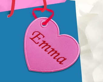 May include: A pink heart-shaped tag with the name "Emma" embroidered in red script. The tag is attached to a blue gift bag with red ribbon handles. The tag has a stitched border.