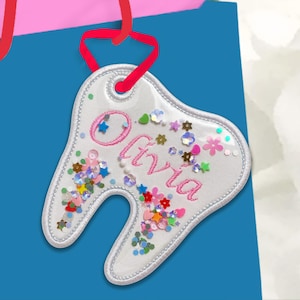 May include: A white tooth-shaped ornament with the name "Olivia" embroidered in pink. The ornament is filled with colorful sequins and small star-shaped confetti. It hangs from a red ribbon against a blue background.