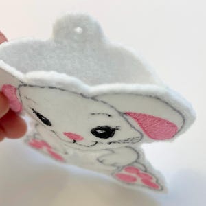 May include: White felt bunny ornament with pink ears and black eyes. The bunny is holding a small pink heart in its paws.