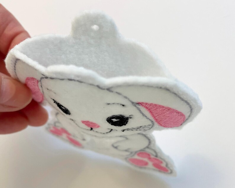 May include: White felt bunny ornament with pink ears and black eyes. The bunny is holding a small pink heart in its paws.