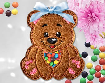 May include: A brown felt teddy bear ornament featuring a clear plastic belly window filled with colorful candy-coated chocolates. The bear has pink stitched paw pads, black embroidered facial features, and a light blue satin ribbon bow on top.