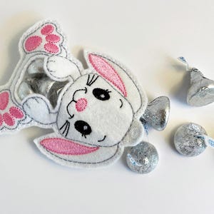 May include: A white felt bunny with pink ears and pink paw prints. The bunny is holding a silver foil wrapped chocolate candy.