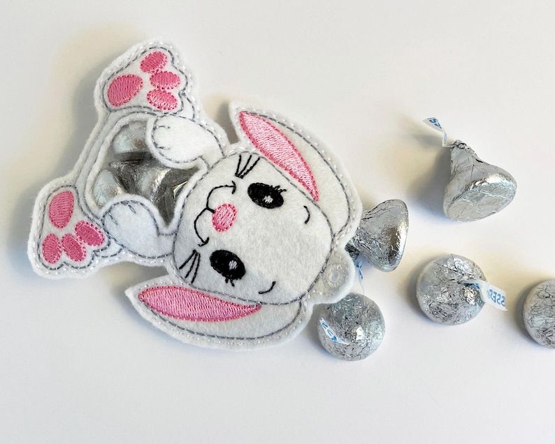 May include: A white felt bunny with pink ears and pink paw prints. The bunny is holding a silver foil wrapped chocolate candy.