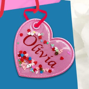 May include: A pink heart-shaped tag with the name "Olivia" in red script. The tag is filled with colorful heart-shaped confetti and has a red ribbon for hanging. The tag is set against a blue background.