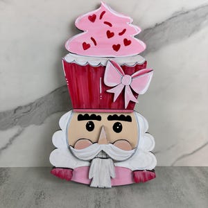 May include: A pink and red wooden nutcracker with a cupcake hat. The cupcake has pink frosting and red hearts. The nutcracker has a white beard and mustache.
