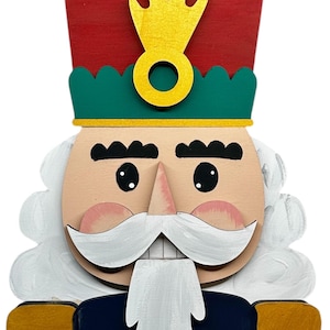 May include: A wooden Christmas nutcracker decoration. The nutcracker has a red hat with green trim, a yellow crown, and a white beard. The uniform is navy blue with gold accents. The face is light tan with pink cheeks.
