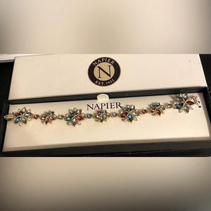 Puede incluir: Una pulsera dorada con detalles en forma de flor, con piedras preciosas rojas, azules y verdes. La pulsera se muestra en una caja blanca con el logotipo "Napier Est. 1922".