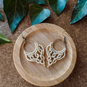 以下が含まれることがあります： 金色のアクセントが付いた白いマクラメのイヤリングのペア。イヤリングは木製のプレートにかけられています。