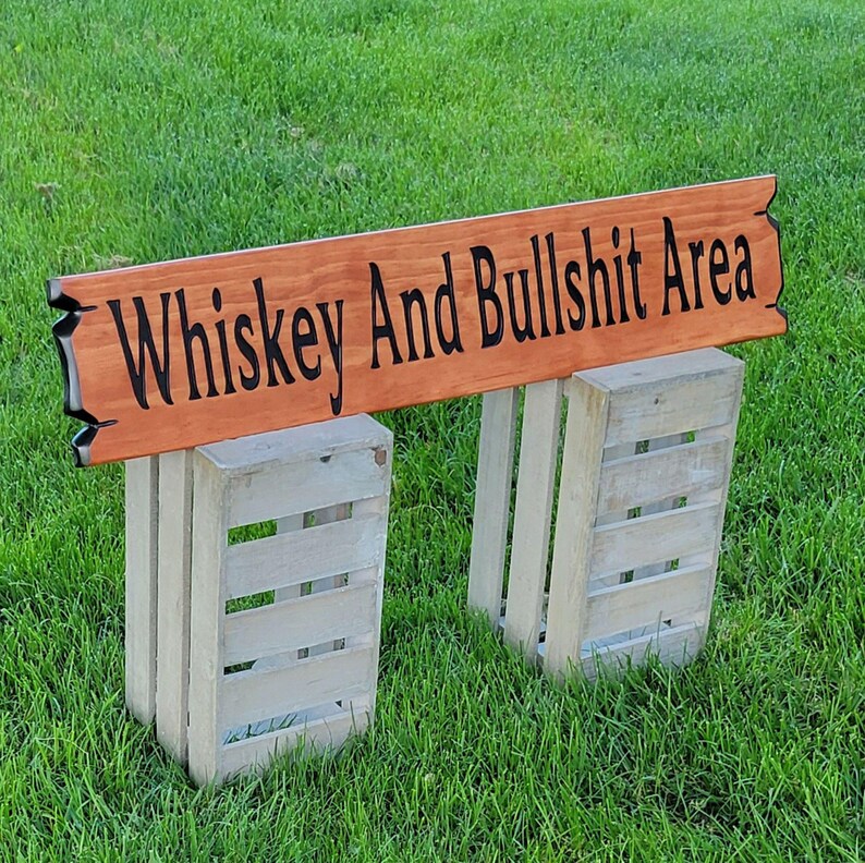 This is a hand carved wooden sign that features "Whiskey and Bullshit Area" Hand carved in the front middle, the saying is hand carved then painted in black. with both ends having a broken board effect, also painted in black. this wooden sign features a gunstock-stained background but you choose your stain color from the drop-down menu. the item is displayed outside on 2 wooden crates to show height and depth.