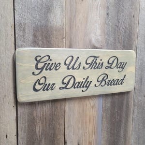 A hand carved wooden sign featuring “Give Us This Day Our Daily Bread” hand carved and painted in black Cursive Lettering with a black painted border trim. Shown in a Classic Gray stain background with a smooth rounded corner. Displayed on a weathered gray barn wood backdrop. Available in 3 sizes and your choice of Rounded or squared corners and stain color.