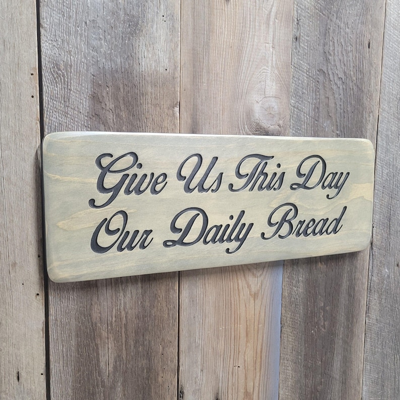 A hand carved wooden sign featuring “Give Us This Day Our Daily Bread” hand carved and painted in black Cursive Lettering with a black painted border trim. Shown in a Classic Gray stain background with a smooth rounded corner. Displayed on a weathered gray barn wood backdrop. Available in 3 sizes and your choice of Rounded or squared corners and stain color.