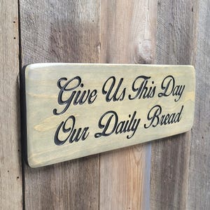 A hand carved wooden sign featuring “Give Us This Day Our Daily Bread” hand carved and painted in black Cursive Lettering with a black painted border trim. Shown in a Classic Gray stain background with a smooth rounded corner. Displayed on a weathered gray barn wood backdrop. Available in 3 sizes and your choice of Rounded or squared corners and stain color.