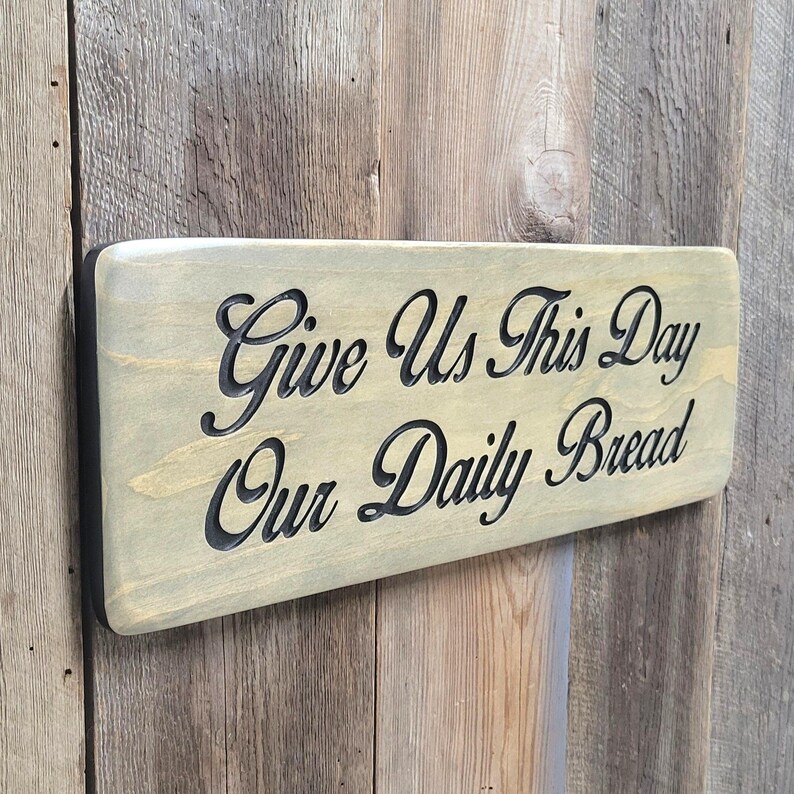 A hand carved wooden sign featuring “Give Us This Day Our Daily Bread” hand carved and painted in black Cursive Lettering with a black painted border trim. Shown in a Classic Gray stain background with a smooth rounded corner. Displayed on a weathered gray barn wood backdrop. Available in 3 sizes and your choice of Rounded or squared corners and stain color.