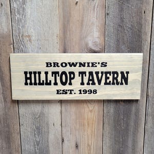 This hand carved wooden sign features a family name and the name of their tavern or bar. with an Est date. you choose our most popular wording of Man Cave, Tavern, Home Bar or you can write your own up to 6 total words, 2 words per line and up to 3 lines. you get your choice of stain color. the hand carved wording is carved then painted in black with black border trim. the Photo'd sign is displayed on an old weather barn wood backdrop.