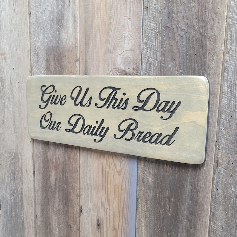 A hand carved wooden sign featuring “Give Us This Day Our Daily Bread” hand carved and painted in black Cursive Lettering with a black painted border trim. Shown in a Classic Gray stain background with a smooth rounded corner. Displayed on a weathered gray barn wood backdrop. Available in 3 sizes and your choice of Rounded or squared corners and stain color.