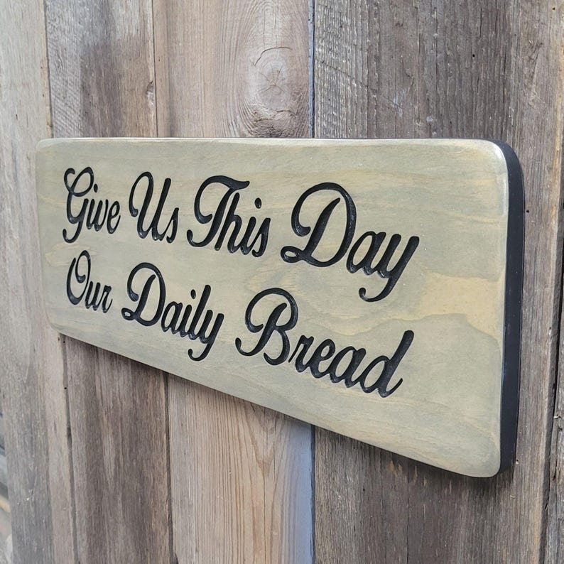 A hand carved wooden sign featuring “Give Us This Day Our Daily Bread” hand carved and painted in black Cursive Lettering with a black painted border trim. Shown in a Classic Gray stain background with a smooth rounded corner. Displayed on a weathered gray barn wood backdrop. Available in 3 sizes and your choice of Rounded or squared corners and stain color.