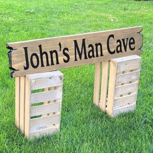 This is a hand carved wooden sign that is custom personalized to your first or last name and says man cave. can be personalized up to 5 words. the wording is carved and painted in black with a broken board effect on both ends on a stained background of your choice from the drop-down menu.