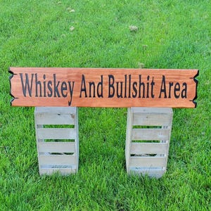 This is a hand carved wooden sign that features "Whiskey and Bullshit Area" Hand carved in the front middle, the saying is hand carved then painted in black. with both ends having a broken board effect, also painted in black. this wooden sign features a gunstock-stained background, but you choose your stain color from the drop-down menu. the item is displayed outside on 2 wooden crates to show height and depth.