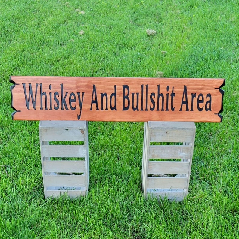This is a hand carved wooden sign that features "Whiskey and Bullshit Area" Hand carved in the front middle, the saying is hand carved then painted in black. with both ends having a broken board effect, also painted in black. this wooden sign features a gunstock-stained background, but you choose your stain color from the drop-down menu. the item is displayed outside on 2 wooden crates to show height and depth.