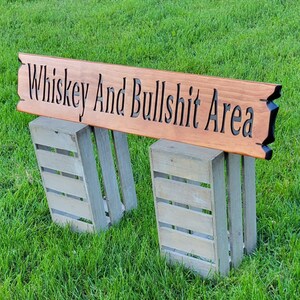 This is a hand carved wooden sign that features "Whiskey and Bullshit Area" Hand carved in the front middle, the saying is hand carved then painted in black. with both ends having a broken board effect, also painted in black. this wooden sign features a gunstock-stained background, but you choose your stain color from the drop-down menu. the item is displayed outside on 2 wooden crates to show height and depth.