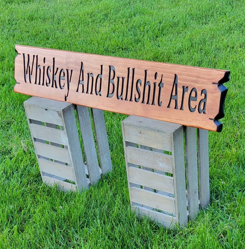 This is a hand carved wooden sign that features "Whiskey and Bullshit Area" Hand carved in the front middle, the saying is hand carved then painted in black. with both ends having a broken board effect, also painted in black. this wooden sign features a gunstock-stained background, but you choose your stain color from the drop-down menu. the item is displayed outside on 2 wooden crates to show height and depth.