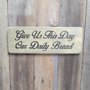 A hand carved wooden sign featuring “Give Us This Day Our Daily Bread” hand carved and painted in black Cursive Lettering with a black painted border trim. Shown in a Classic Gray stain background with a smooth rounded corner. Displayed on a weathered gray barn wood backdrop. Available in 3 sizes and your choice of Rounded or squared corners and stain color.