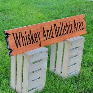 This is a hand carved wooden sign that features "Whiskey and Bullshit Area" Hand carved in the front middle, the saying is hand carved then painted in black. with both ends having a broken board effect, also painted in black. this wooden sign features a gunstock-stained background but you choose your stain color from the drop-down menu. the item is displayed outside on 2 wooden crates to show height and depth.