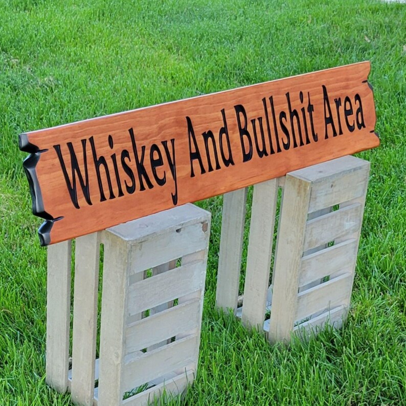 This is a hand carved wooden sign that features "Whiskey and Bullshit Area" Hand carved in the front middle, the saying is hand carved then painted in black. with both ends having a broken board effect, also painted in black. this wooden sign features a gunstock-stained background but you choose your stain color from the drop-down menu. the item is displayed outside on 2 wooden crates to show height and depth.