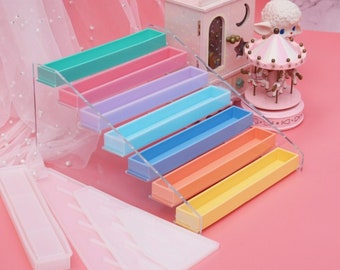 May include: A clear acrylic tiered display stand with six colorful rectangular trays. The trays are in shades of green, pink, purple, blue, light blue, orange, and yellow. The stand is on a pink surface.