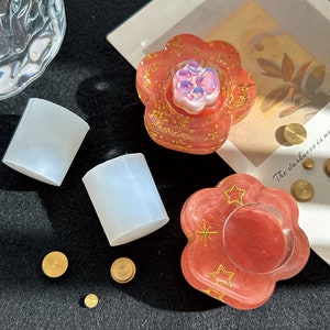 May include: Two white silicone moulds, one shaped like a cylinder and the other shaped like a paw print. A pink resin paw print mould with gold glitter and a star design. A pink resin mould with a star design and a clear centre. The moulds are surrounded by small gold metal circles.
