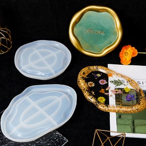 May include: Two clear silicone molds for resin casting in the shape of a wavy oval with four divided sections. The molds are on a black surface. A finished resin tray with gold leaf and dried flowers is on a magazine. A gold tray with a green background and the text "Best wishes" is in the background.