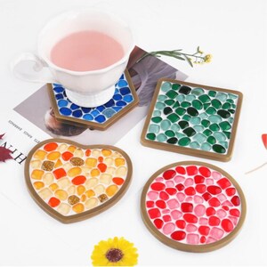 Puede incluir: Una taza blanca con líquido rosa descansa sobre un posavasos de mosaico azul y marrón. Otros posavasos presentan diseños de mosaico verde, rojo y naranja en formas cuadradas, de corazón y redondas.