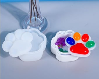 Può includere: Scatola bianca a forma di impronta di zampa con un coperchio. Il coperchio presenta un design a forma di impronta di zampa colorato con accenti rossi, blu, verdi, arancioni e gialli. La scatola è aperta e contiene piccoli oggetti.