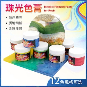 Op de afbeelding: Een verzameling kleine potjes metallic pigmentpasta voor hars. De potjes hebben witte deksels en kleurrijke etiketten. De afbeelding bevat tekst in het Engels en Chinees, en een decoratief harskunstwerk.