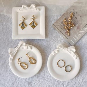Puede incluir: Bandejas de cerámica blanca para joyas con detalles de lazo. Las bandejas contienen pendientes de oro, anillos y una rama dorada decorativa. Una bandeja es rectangular y dos son redondas. Los pendientes tienen detalles de perlas.
