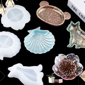 May include: Six different silicone molds for resin casting. The molds are shaped like a seashell, a flower, a bear, a cat, and two other abstract shapes. The molds are all white and have a smooth surface.