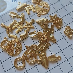 Peut inclure: Un assortiment de breloques et de pendentifs dorés de formes variées, notamment des étoiles, des nuages, des pièces de monnaie et des motifs abstraits. Ces petites pièces détaillées sont probablement destinées à la fabrication de bijoux ou à l'artisanat.
