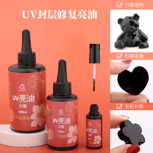 Peut inclure: Trois flacons de résine UV, étiquetés "UV 亮油" (UV Bright Oil) en caractères chinois, de différentes tailles : 200 ml, 100 ml et 15 ml. Les flacons sont sur un fond rose. L’image montre également trois exemples d’objets en résine : un ours noir, un cœur noir et une fleur noire.
