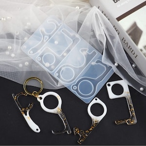 May include: A silicone mold with five different shapes for making keychains. The mold is clear and shows the shapes of the keychains. There are three finished keychains made of white resin with gold glitter. The keychains are shaped like a door handle, a bottle opener, and a key.