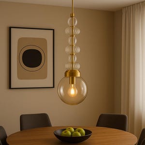 May include: A modern pendant light fixture with a gold finish and clear glass globes. The light hangs above a wooden dining table with a bowl of green apples. The room has a neutral color palette.